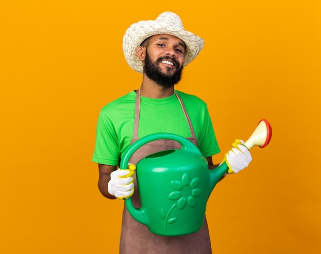 Улыбающийся молодой афро-американский парень садовник в садовой шляпе и перчатках держит лейку