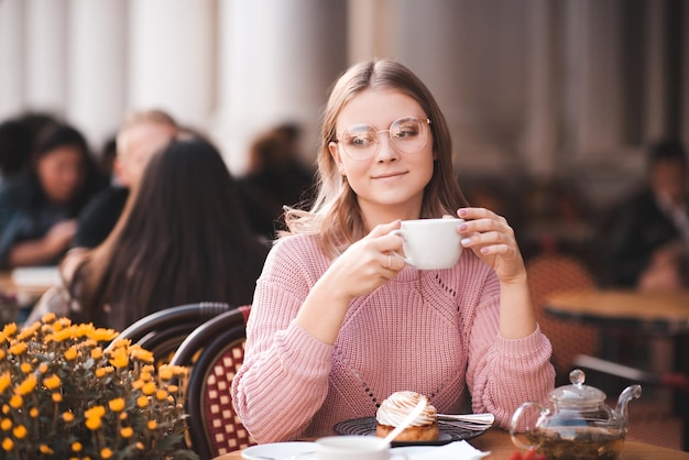 Smiling young girl drinking tea and eating cake in cafe on street