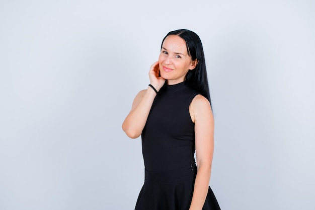 Smiling young girl is looking at camera by holding hand near cheek on white background