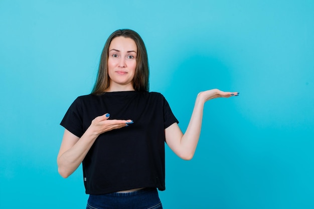 Photo smiling young girl is pointing right with hands on blue background