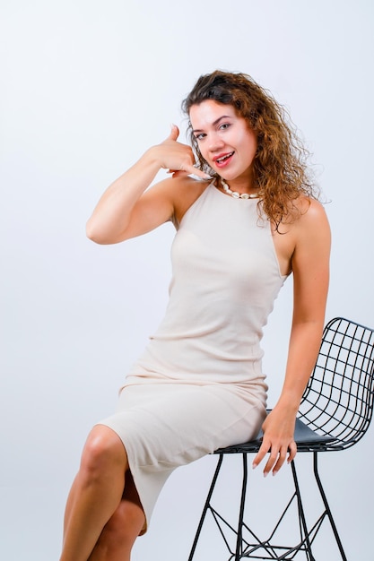 Smiling young girl is showing phone gesture with hand by sitting on white background