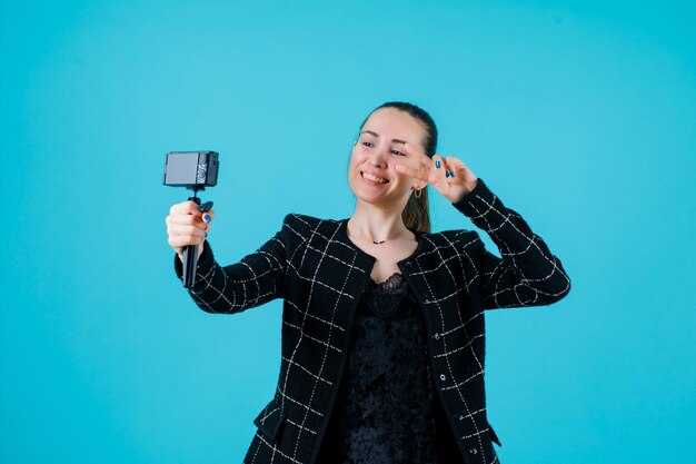 Smiling young girl is taking selfie with mini camera by showing two gesture on blue background