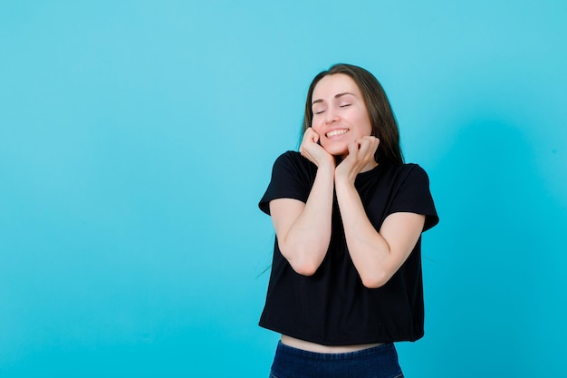 Smiling young girl is wishing by holding hands on cheeks on blue background