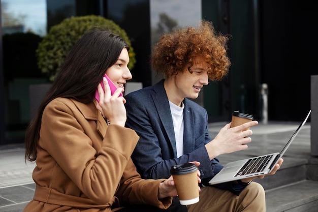 Улыбающаяся молодая девушка и мужчина сидят возле современного офиса Привлекательная женщина разговаривает по смартфону