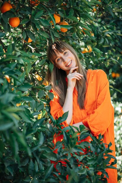 Smiling young girl in orange dress is posing to camera by hodling forefinger on chin in orange garden