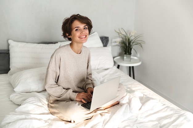 Smiling young girl relaxing in bed in the morning, using laptop computer