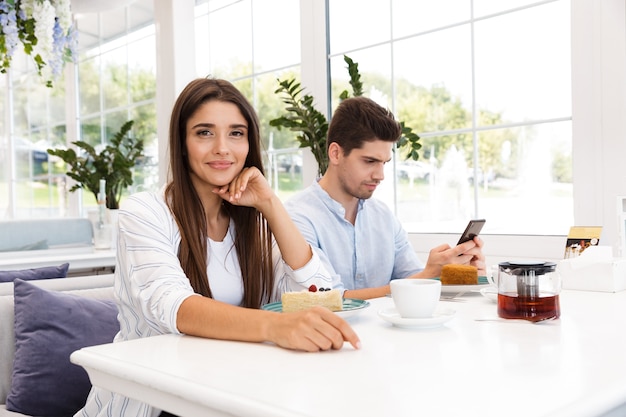 Smiling young girl sitting at the cafe table while her boyfriend using mobile phone