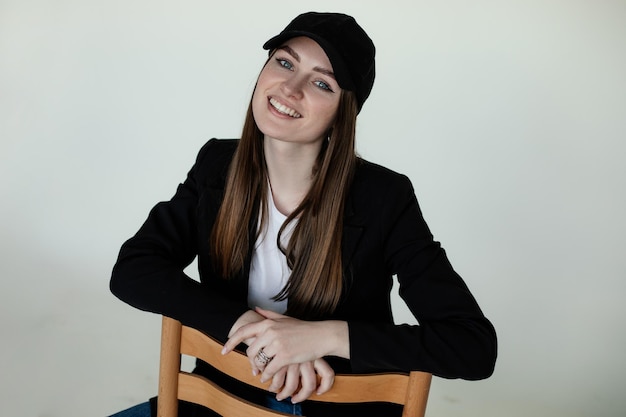 Smiling young girl sitting on chair gray studio background copy space Female casual black blazer and cap trendy clothes and headdresses Monochromatic collection of fashionable clothing for women