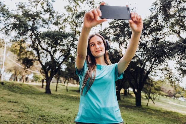 Smiling young girl using headphones, taking a selfie with her mobile in a natural space