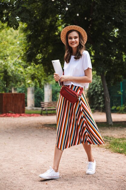 Smiling young girl walking with laptop computer at the park outdoors