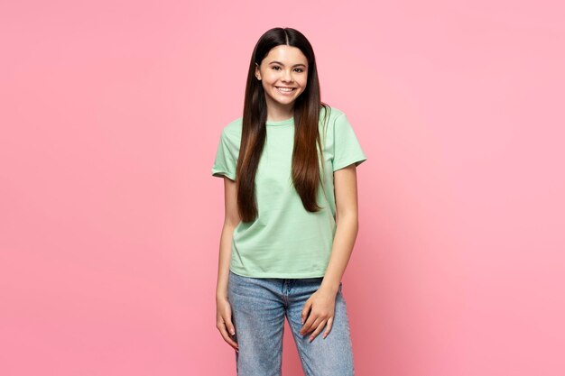 Photo smiling young girl wearing green t shirt and jeans looking at camera isolated on pink background