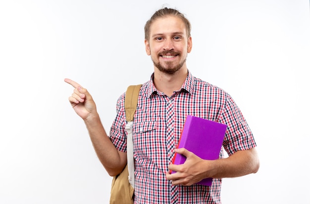 Smiling young guy student wearing backpack holding books points at side 