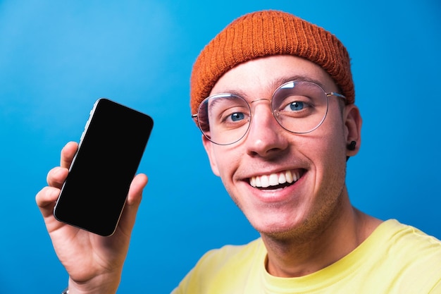 Photo smiling young guy with glasses showing smartphone app with clean screen for mockup