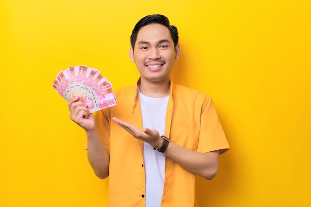 Smiling young handsome asian man showing money banknotes isolated on yellow background