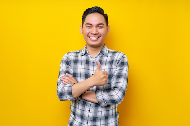 Smiling young handsome Asian man wearing casual clothes showing thumbs up gesture Approving expression looking at the camera isolated on yellow background People lifestyle concept