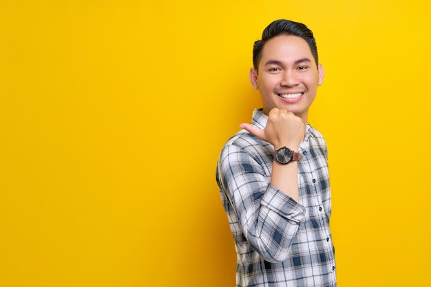 Smiling young handsome Asian man wearing a plaid shirt pointing thumb aside at blank space for text isolated on yellow background People lifestyle concept