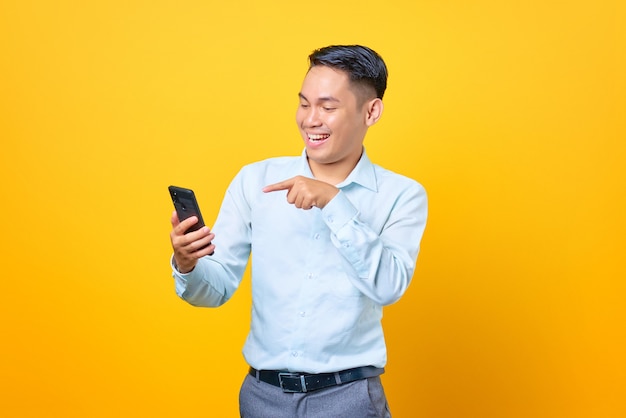 Smiling young handsome businessman pointing at smartphone on yellow background