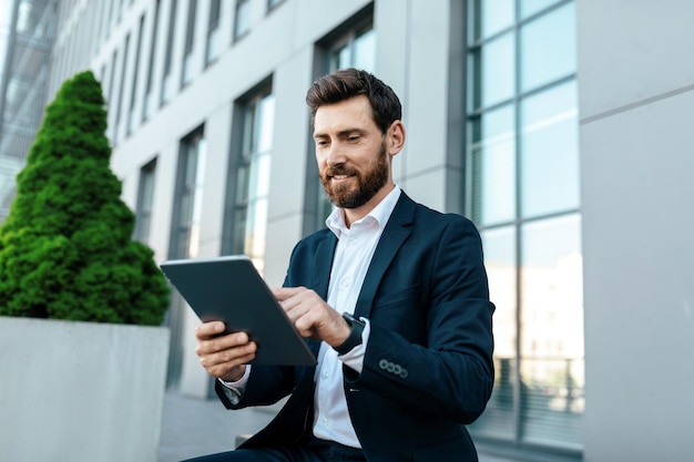 Smiling young handsome european businessman with beard in suit typing on tablet near modern office