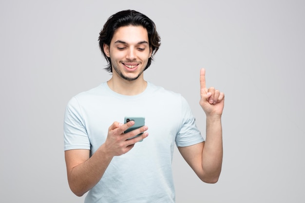 smiling young handsome man holding mobile phone looking at it pointing up isolated on white background