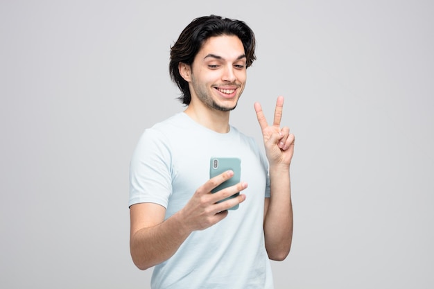 smiling young handsome man holding mobile phone showing peace sign taking selfie isolated on white background