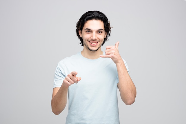 Smiling young handsome man looking and pointing at camera showing call gesture isolated on white background