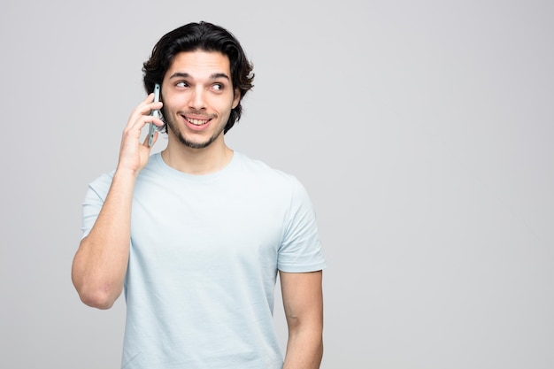 smiling young handsome man looking at side while talking on phone isolated on white background with copy space