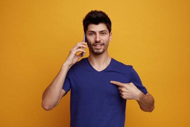 Smiling young handsome man talking on phone looking at camera pointing at himself on yellow background