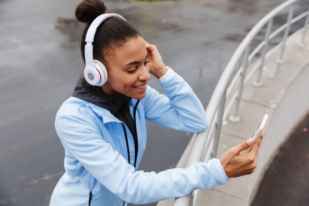 笑顔の若い健康なアフリカのスポーツウーマンがスタジアムで無線イヤホンを使って音楽を聴き、携帯電話を持って、レールに寄りかかって