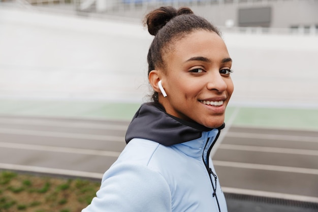 smiling young healthy african sportswoman listening to music with wireless earphones wrking out at the stadium