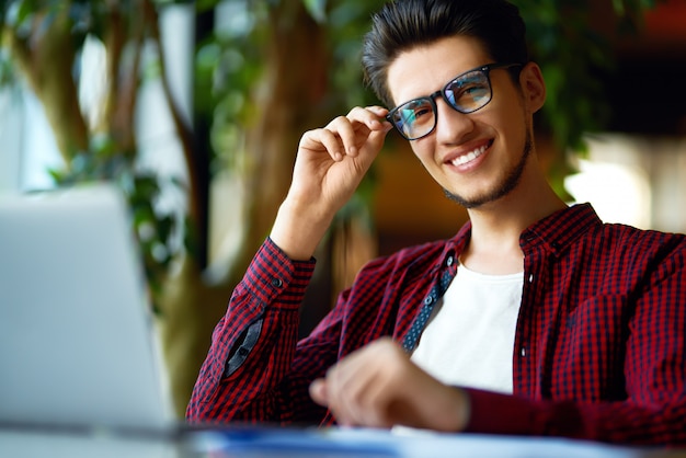 テーブルの上のラップトップでメガネの流行に敏感な若い男の笑みを浮かべてください。プログラマー、Web開発者、オフィスで働いているデザイナーがモバイルとデスクトップのWebサイトのバージョンを比較します。