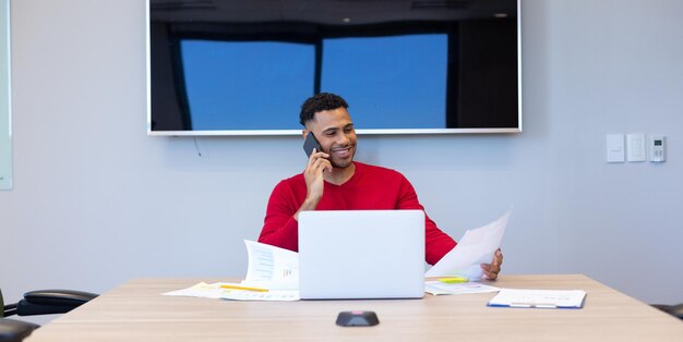 Smiling young hispanic businessman talking through smartphone while analyzing document in boardroom