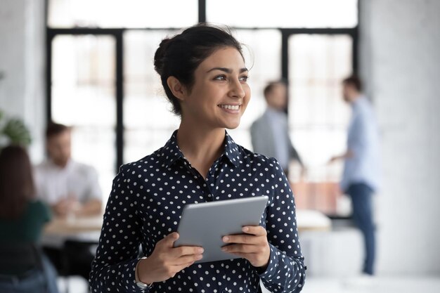 Smiling young Indian female employee hold tablet look in distance thinking happy millennial biracial woman worker distracted from pad gadget lost in thoughts visualizing business vision concept