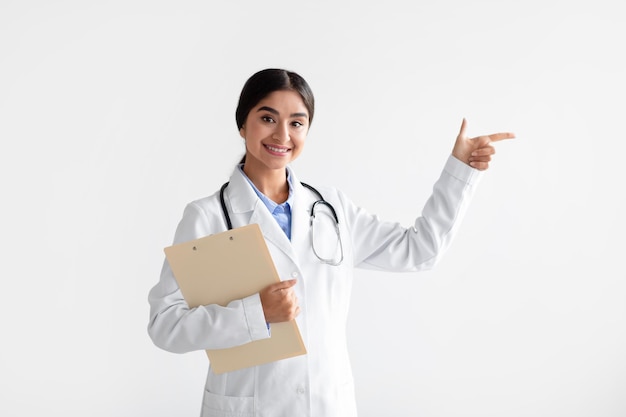 Smiling young indian lady in white coat with tablet shows finger at empty space isolated on white background