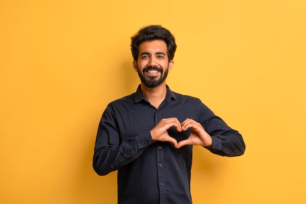 Smiling young indian man showing heart gesture with hands near chest