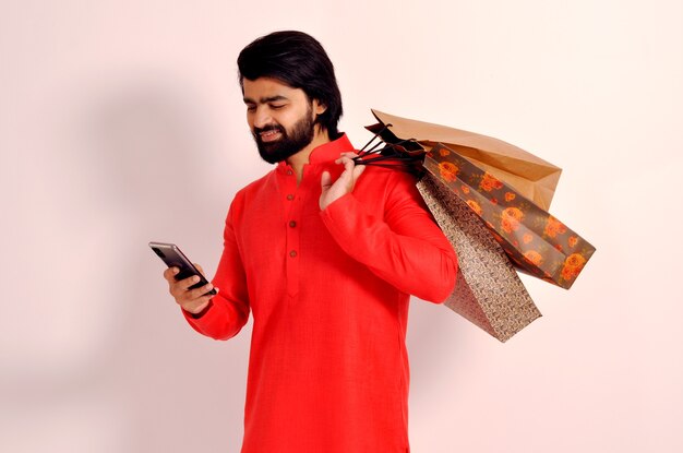 Sorridente giovane indiano uomo che indossa kurta tenendo le borse della spesa e guardando il telefono cellulare