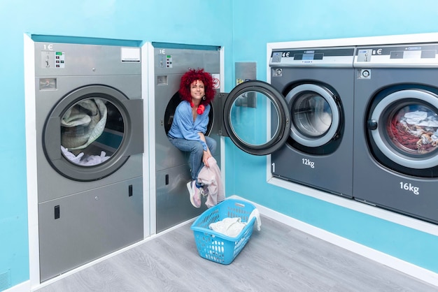 A smiling young latin woman with afro hair sitting inside a washing machine in a blue automatic laundry room