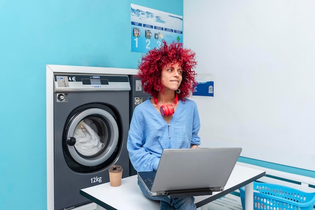 A smiling young latin woman with red afro hair sits at the table in the blue laundry room working on her laptop while waiting for the laundry to be done her red headphones around her neck