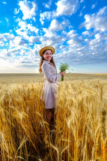 Smiling young long-haired woman in straw hat smiling holds a wild flower bouquet in wheat field at dawn. Model looks direct at camera, vertical orientation