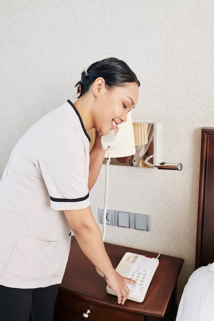 Smiling young maid making phone call to hotel reception to inform that she cleaned the room for the guests