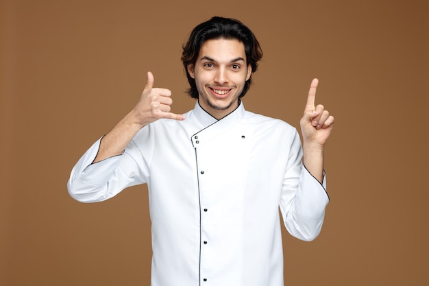 smiling young male chef wearing uniform looking at camera pointing up showing call gesture isolated on brown background