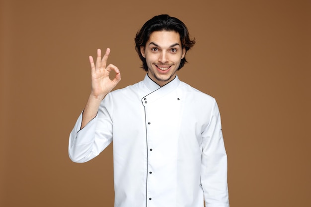 Sorridente giovane chef maschio che indossa l'uniforme guardando la fotocamera che mostra segno ok isolato su sfondo marrone