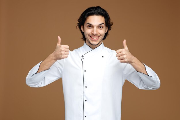 smiling young male chef wearing uniform looking at camera showing thumbs up isolated on brown background