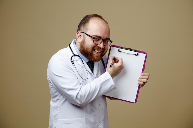 Sorridente giovane medico maschio con occhiali camice da laboratorio e stetoscopio intorno al collo che mostra appunti che punta la penna guardando la fotocamera isolata su sfondo verde oliva