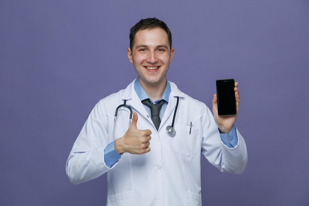 Sorridente giovane medico maschio che indossa un abito medico e uno stetoscopio intorno al collo guardando la fotocamera che mostra il telefono cellulare e il pollice in su isolato su sfondo viola