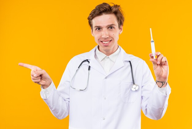 Smiling young male doctor wearing medical robe with stethoscope holding syringe points at side