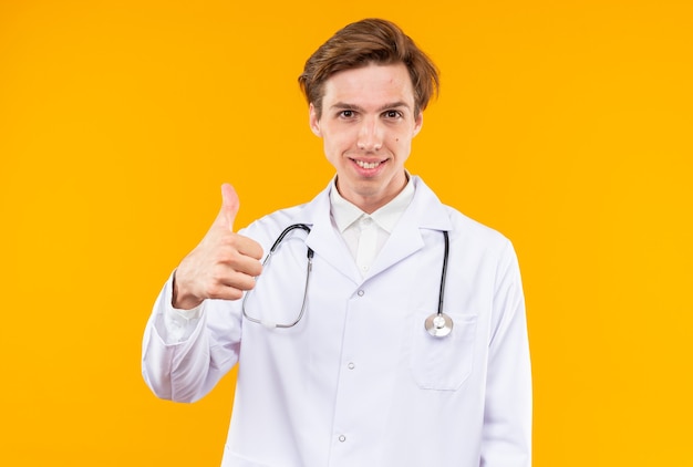 Smiling young male doctor wearing medical robe with stethoscope showing thumb up isolated on orange wall