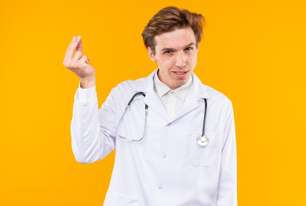 Smiling young male doctor wearing medical robe with stethoscope showing tip gesture isolated on orange wall