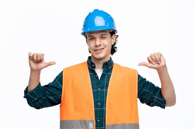 Smiling young male engineer wearing safety helmet and safety vest looking at camera pointing at himself winking isolated on white background