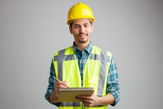 Smiling young male engineer wearing safety helmet and uniform looking at camera holding pencil and note pad isolated on white background