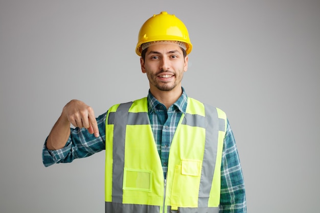 Smiling young male engineer wearing safety helmet and uniform looking at camera pointing down isolated on white background